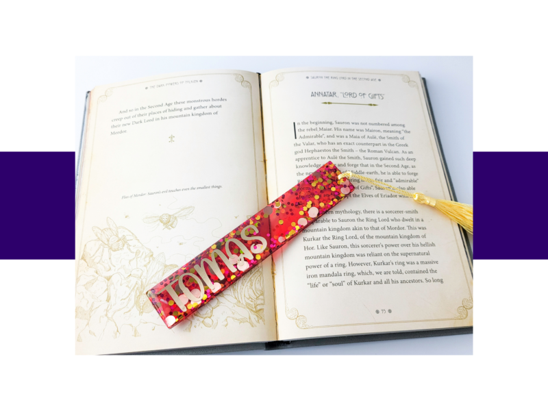 Open book with decorative page borders and visible text, a glittery pink confetti bookmark rests diagonally across the pages.
