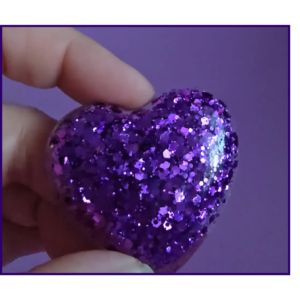Close-up of a finger pinching a purple glittery ball against a purple background and lighting that highlights the sparkles.
