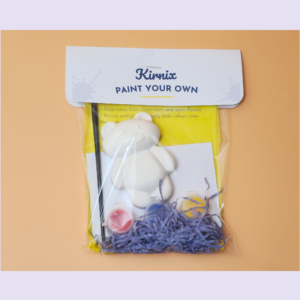 Kit titled 'Kirnix Paint Your Own' containing a white bear figure and painting supplies in a yellow bag on an orange background