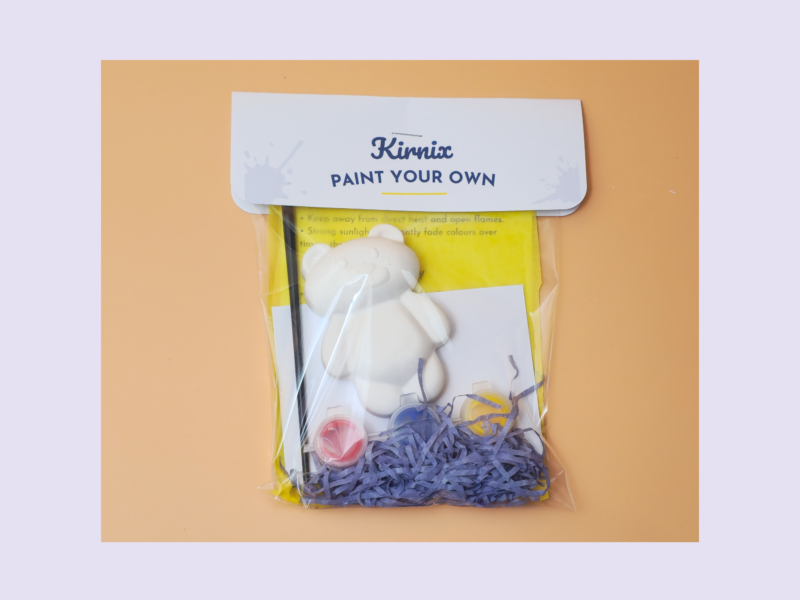 KNP-PYO-BEARS-mini_011. Hero photo Kit titled 'Kirnix Paint Your Own' containing a white bear figure and painting supplies in a yellow bag on an orange background