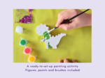 Person uses a paintbrush to color a white dinosaur cutout with green paint, surrounded by paints and glitter on a yellow craft table.