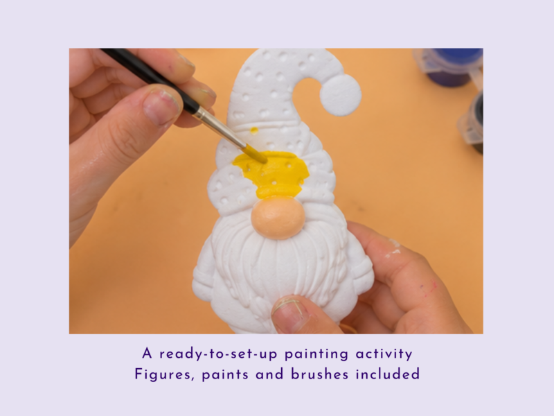 Hands paint a white gnome figurine with yellow on an orange surface, using a brush.
