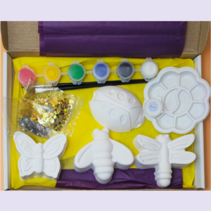 Open craft kit showing butterfly-shaped molds, small paint pots in yellow, green, blue, gray and white, glitter bag, and purple/yellow tissue paper in a box.