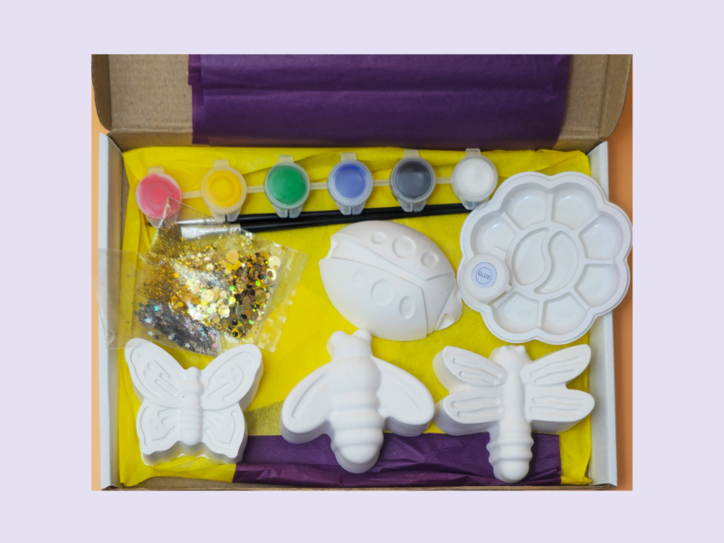Open craft kit showing butterfly-shaped molds, small paint pots in yellow, green, blue, gray and white, glitter bag, and purple/yellow tissue paper in a box.