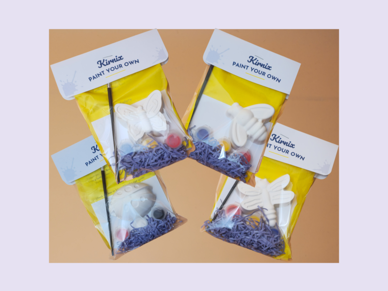 DIY paint kits with yellow packaging and white butterfly shapes, scattered paint pots and brushes on a purple backdrop