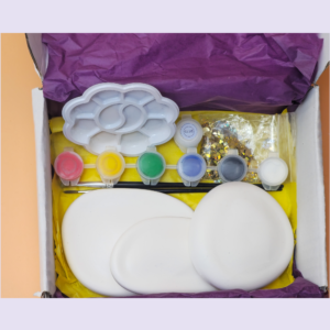 Open box of art supplies with paint pots, a white cloud-shaped palette, glitter, and tissue paper assembly in a yellow tray.
