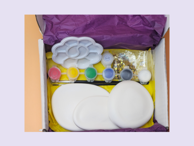 KNP-PYO-STONES-0011. Hero photo Open box of art supplies with paint pots, a white cloud-shaped palette, glitter, and tissue paper assembly in a yellow tray.