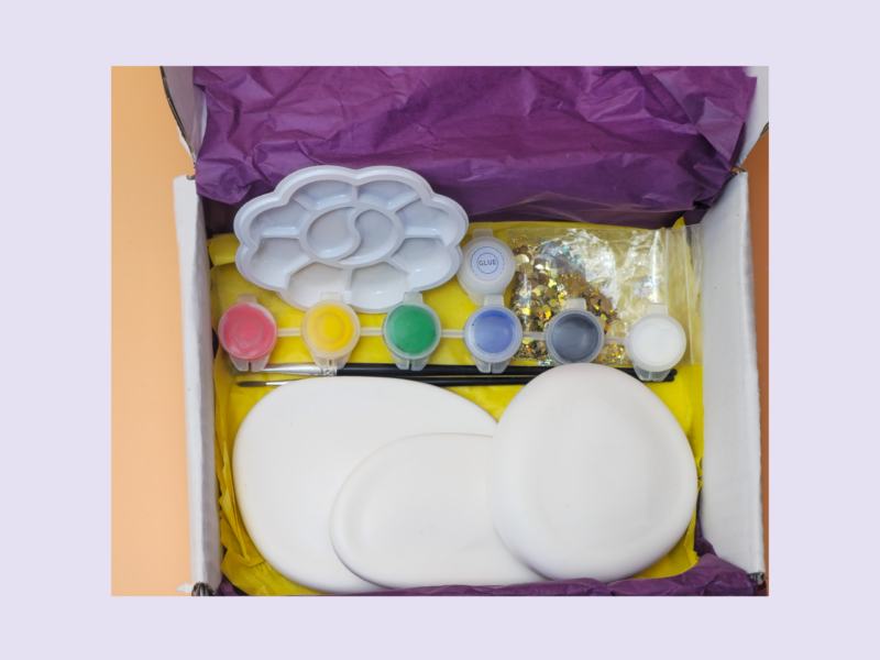Open box of art supplies with paint pots, a white cloud-shaped palette, glitter, and tissue paper assembly in a yellow tray.