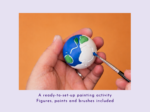 Close-up of a hand holding a small globe being painted with blue paint, orange background behind.
