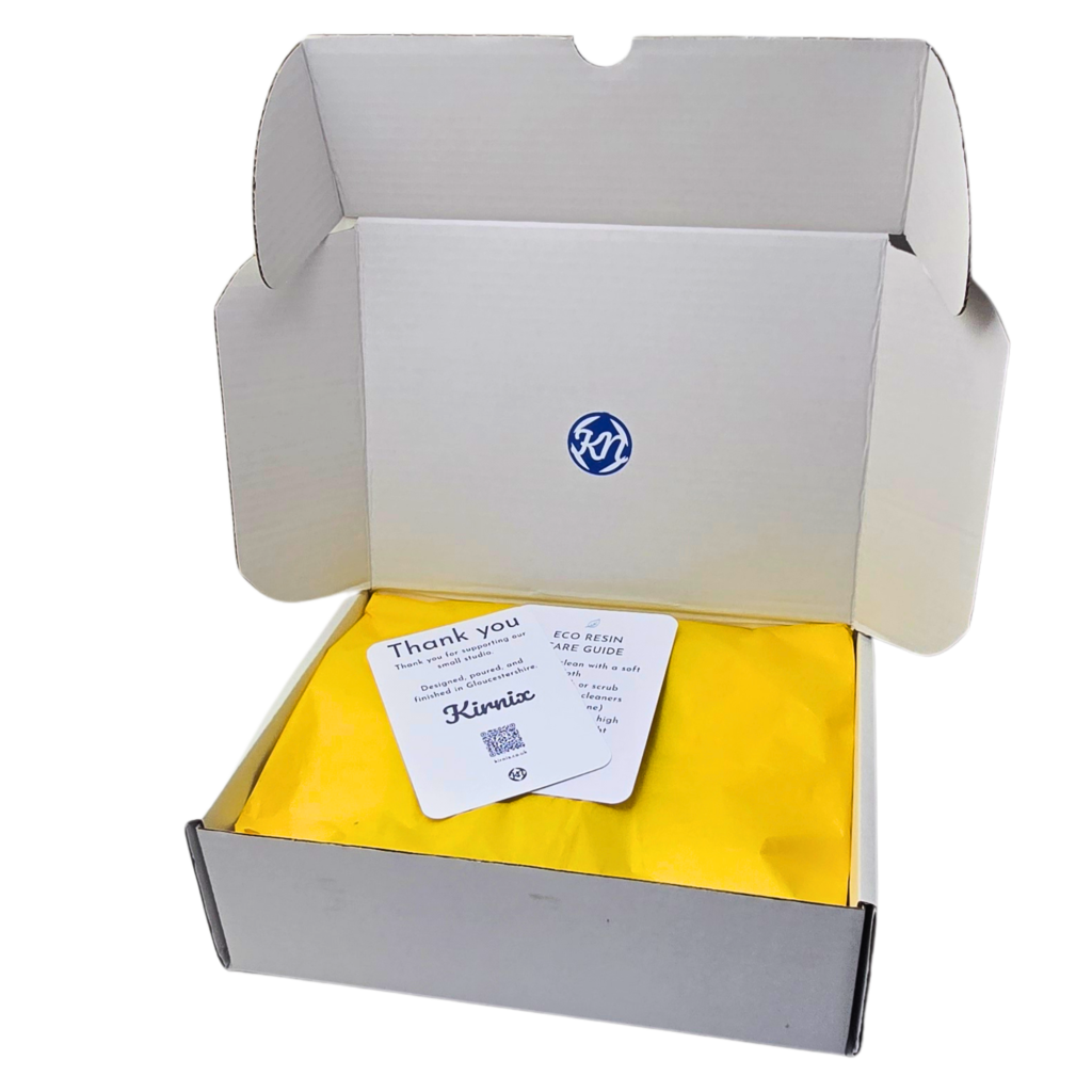 Open white shipping box with yellow packing paper and a small 'Thank you' card on top. A blue logo is visible inside the box.