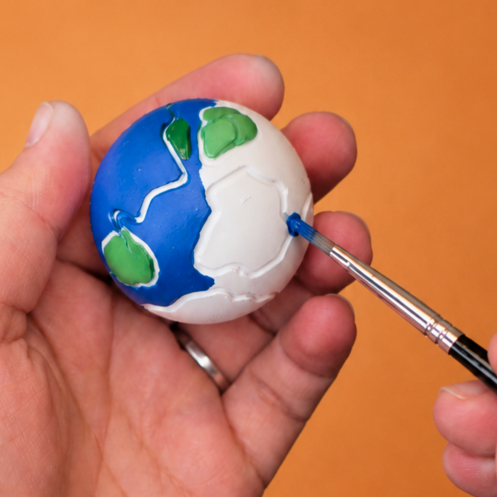 Close-up of a hand painting a small globe with blue oceans and green continents against an orange background.