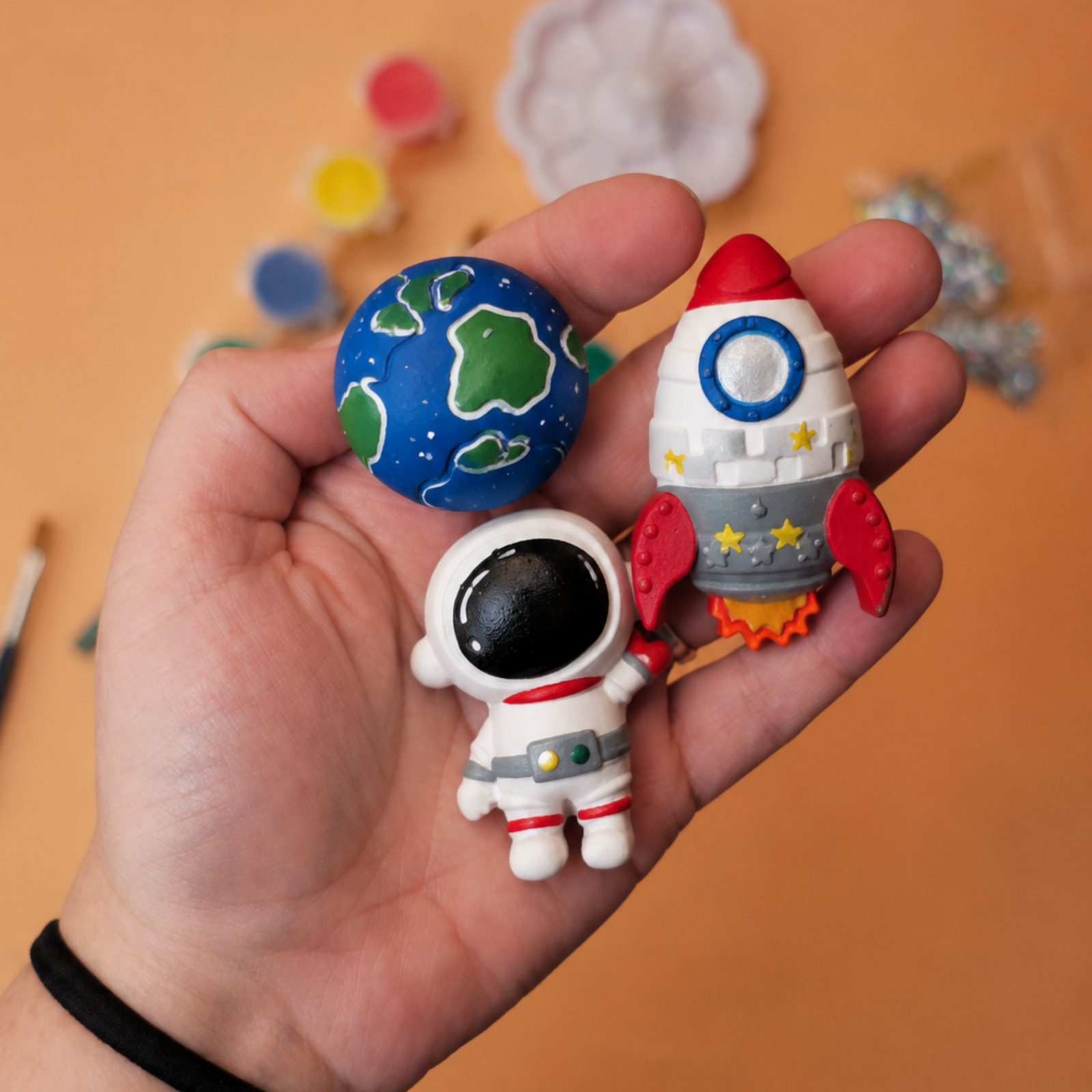 Hand holding a small space-themed toy rocket and a blue planet egg against an orange background.