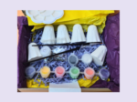 Six unpainted white eco resin ghost figures in an orange gift box with blue shredded tissue and a Kirnix paint your own label, photographed on an orange background