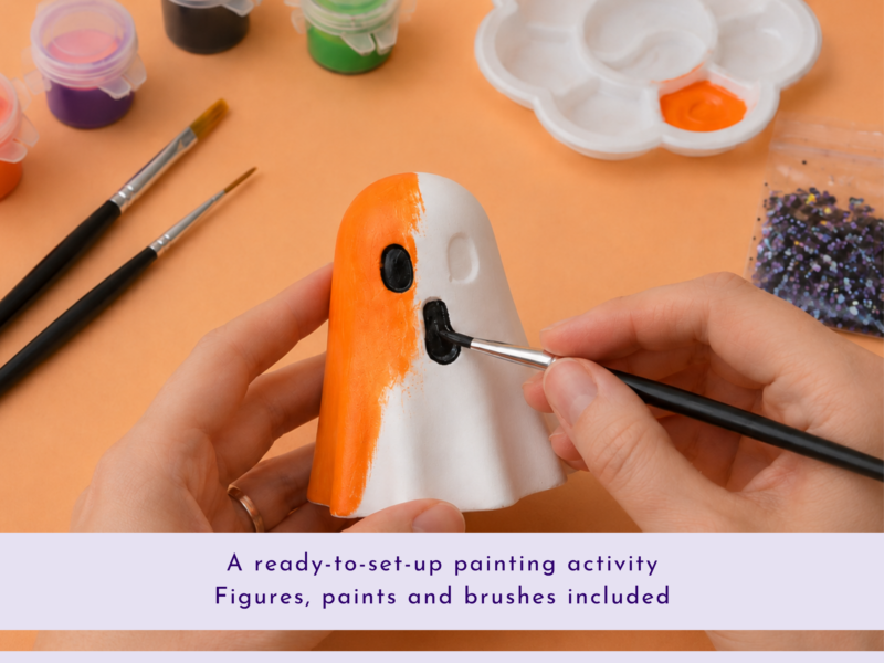 Hands painting an orange-and-white figurine with brushes, surrounded by paint pots and a palette on an orange workspace