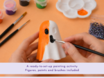 Hands painting an orange-and-white figurine with brushes, surrounded by paint pots and a palette on an orange workspace