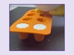 Close-up of an orange silicone mold with circular cavities being pressed by a yellow tool or finger, on a dark surface.