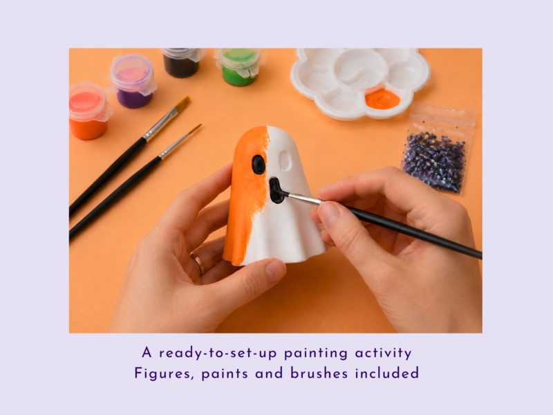 Hands painting a small white-and-orange figurine with brushes and paint supplies nearby on an orange background.