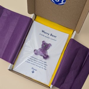 Inside a cardboard box, a children's book titled 'Worry Bear: A little pocket comfort' rests with purple tissue paper nearby.
